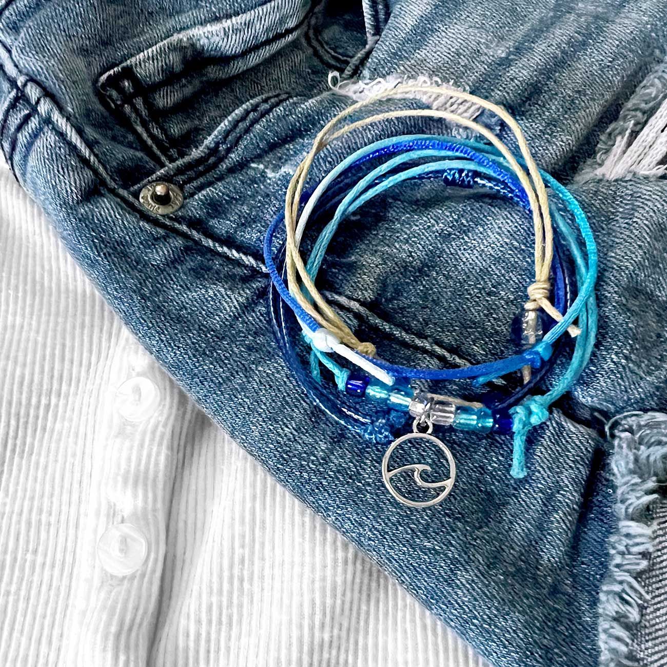 wave bracelet pack with blue and beige cords, glass beads and silver-tone wave charm on faded denim beside a white shirt.