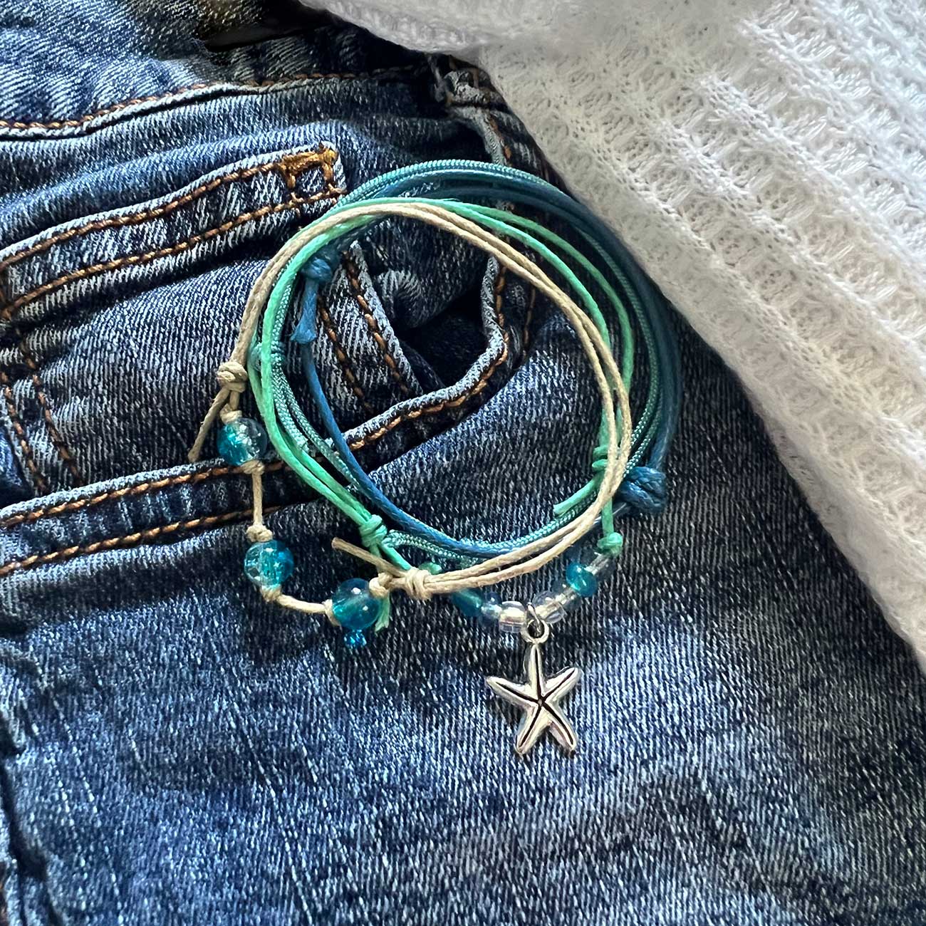 starfish bracelet stack with turquoise cords, blue glass beads and silver-tone starfish charm on denim beside white fabric.