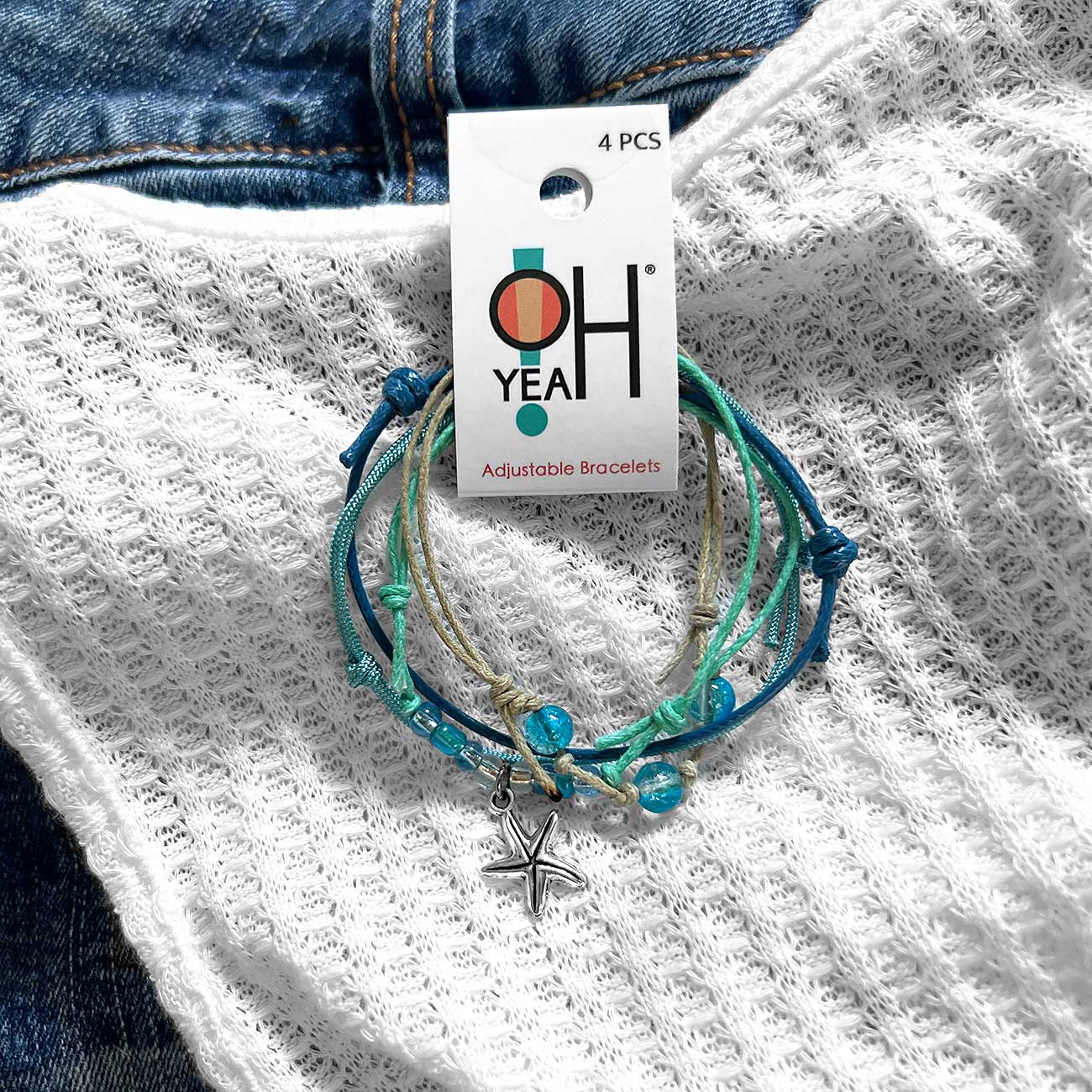 starfish bracelet stack with blue glass beads, silver-tone charm and O Yeah hang tag on white knit fabric over denim.