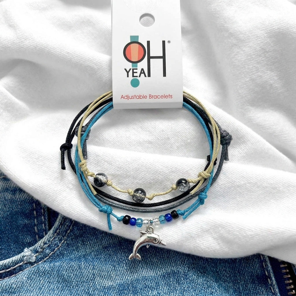 dolphin bracelet charm on a black, blue and beige bracelet stack with O Yeah hang tag, displayed on white fabric and denim.