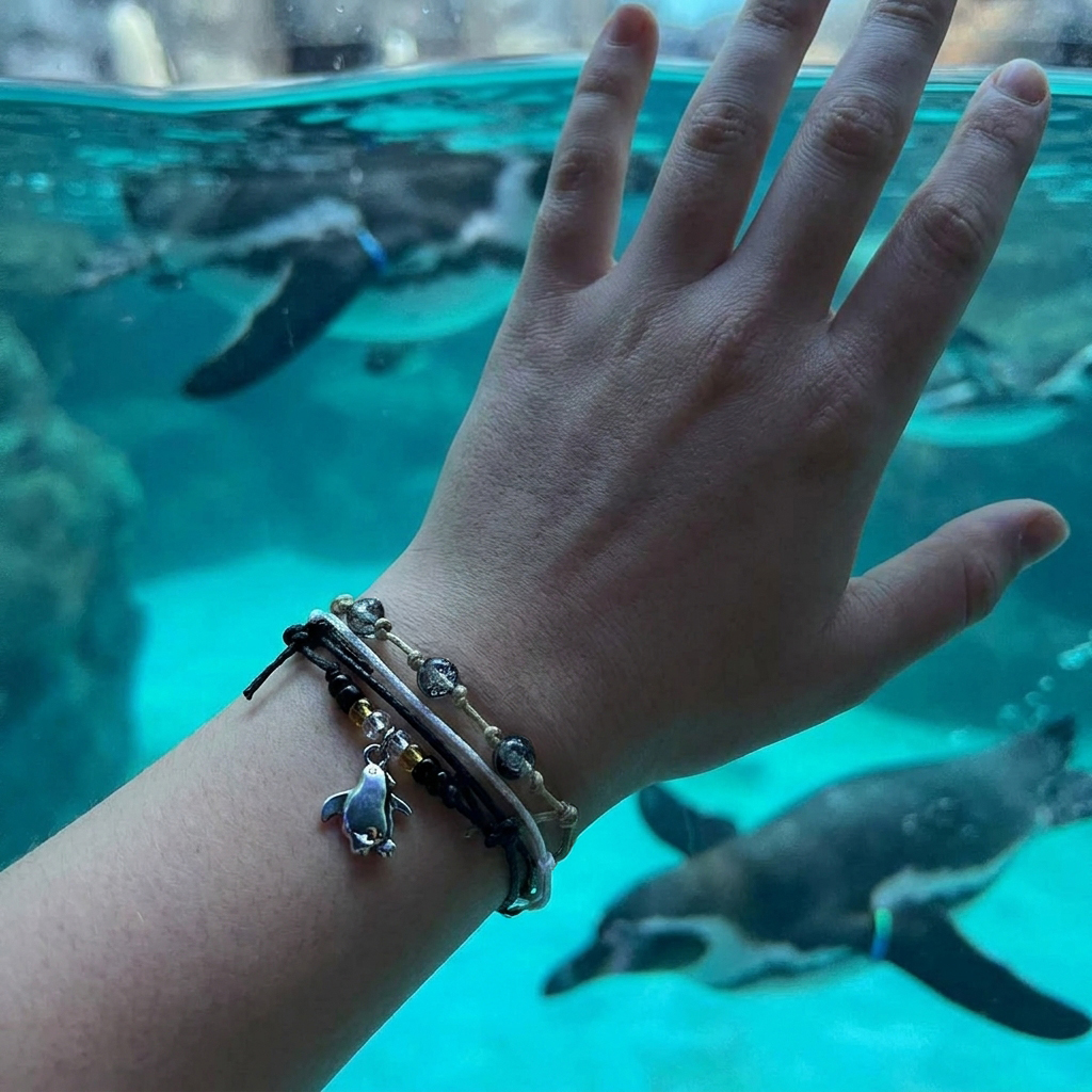 penguin bracelet set on a wrist with black, white and beige cords and silver-tone charm as penguins swim in turquoise water.